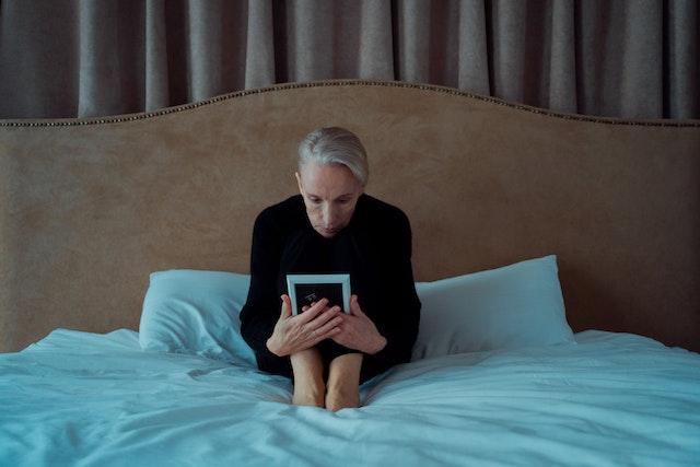 A woman sits on a bed gazing intently at a photo in a white frame.