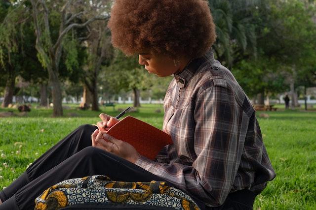 A woman sits in a park thoughtfully writing in a notebook.